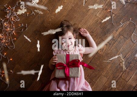 Little happy girl in a pink dress lies on the wooden floor. Holds a Christmas box in craft packaging, a Christmas gift with a red ribbon. Snowflakes h Stock Photo