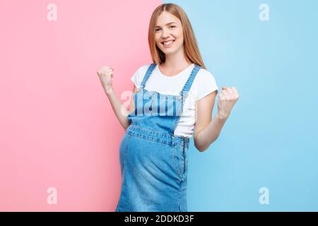 A happy confident young beautiful pregnant woman in a denim jumpsuit shows a winning gesture while standing on an isolated pink and blue background Stock Photo