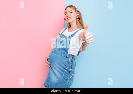 Happy cute beautiful young cheerful pregnant woman enjoys listening to music with headphones while standing on an isolated pink and blue background Stock Photo