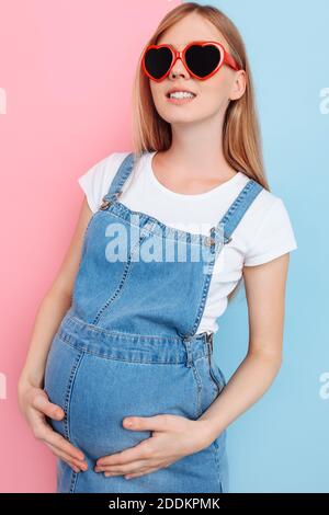 Emotional happy young pregnant woman in denim overalls, wearing heart-shaped glasses, posing standing on an isolated pink and blue background Stock Photo