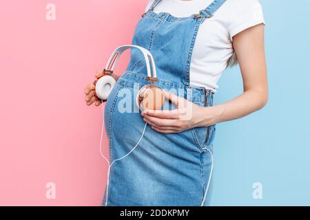 Close - up of a pregnant woman's belly holding headphones with music near her belly, on a pink and blue background Stock Photo