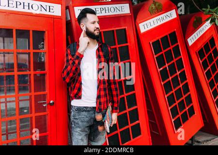 Stylish man with a beard with wireless headphones, hipster listens to music, standing on the background of a retro telephone box