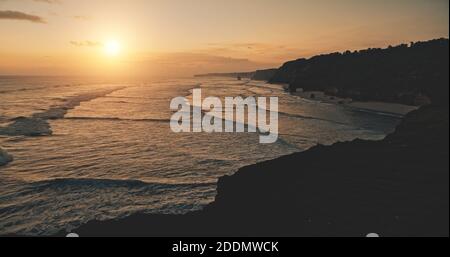 Relax serene seascape sunset silhouette of ocean cliff coast aerial view. Cinematic sun set light at dark rock wall. Amazing sea bay waves water washed sand beach of Bawana, Sumba Island, Indonesia Stock Photo