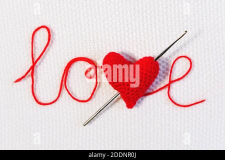 Valentine's day, wedding backdrop. Crochet hook pierced red knitted volume heart like an arrow in word love from threads on white knitted background. Stock Photo
