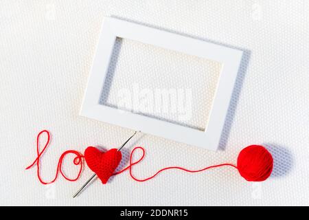Valentine's day or wedding backdrop. Red knitted volume heart in word love, ball of thread and white frame on knitted background. Template for design, Stock Photo