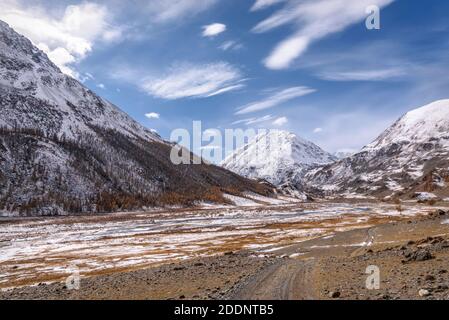 Amazing autumn landscape with a winding dirt road, river, snowy mountains, golden larch trees on the slopes and the first snow against a background of Stock Photo