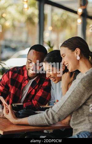 Group of friends sitting at cafe table and looking at mobile phone. Young man and women looking at pictures on smart phone and smiling. Stock Photo