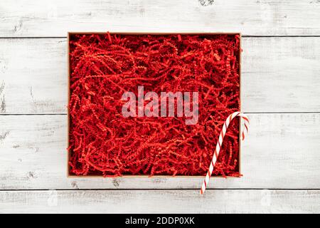 Opened gift box with red shredded paper on white wooden background. Flat lay, top view. Box for your product replacement. Xmas. Stock Photo
