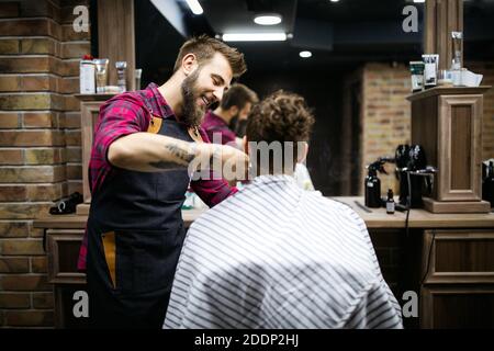 Young bearded man getting haircut by hairdresser with electric razor at barber shop Stock Photo