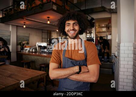 Handsome young male waiter wearing apron standing with crossed arms in cafe looking at camera Stock Photo