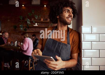 Portrait of a happy waiter standing at restaurant leaning on wall holding digital tablet Stock Photo
