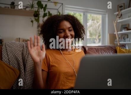 Cheerful african woman relaxing on couch waving hand while on video call using laptop and earphones Stock Photo