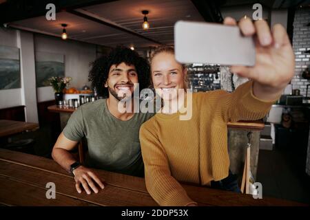 Smiling young woman sitting with male friend taking selfie in cafe using mobile phone Stock Photo