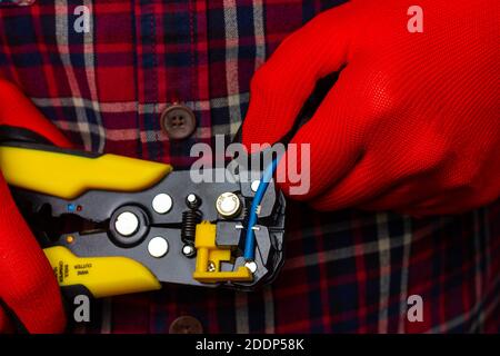 A guy in a plaid shirt and red work gloves is stripping electrical wires with yellow pliers. Stock Photo