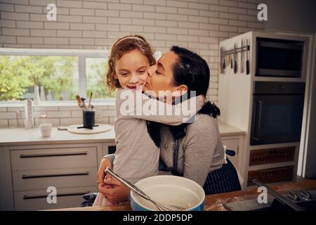 Loving mother embracing daughter and kissing her on cheek while preparing meal on kitchen counter Stock Photo
