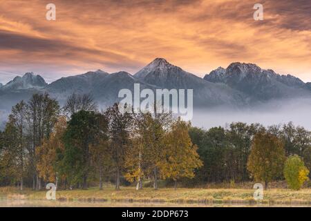 Magical Sunrise over High Tatra Mountains. Dramatic Sky and Autumnal Foliage Colors. Mist and Fog over Lake. Stock Photo