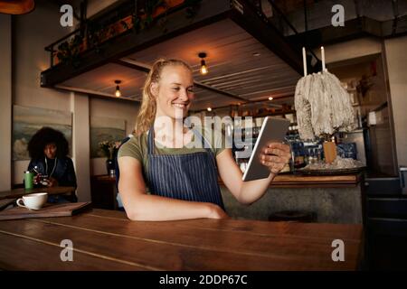 Smiling female waiter standing leaning on table using digital tablet in modern cafe Stock Photo