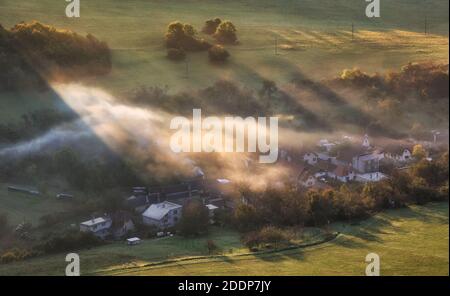 Countryside rural landscape with village over sunbeam in misty morning. Stock Photo