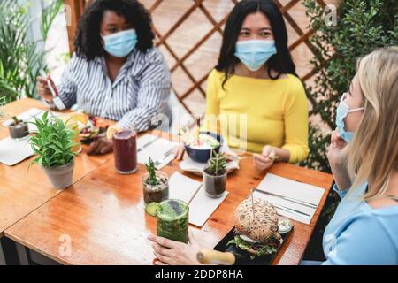 Multiracial young women eating brunch during coronavirus outbreak - Girls wearing protective masks at trendy restaurant - Healthy lifestyle and social Stock Photo