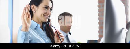 Smiling businesswoman sits at computer screen with microphone and waves hand with colleague in background in office Stock Photo