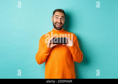 Online shopping. Young man thinking and showing smartphone screen, looking dreamy at upper left corner, standing over light blue background Stock Photo