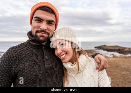 Beautiful young couple in love standing on sea resort looking at camera posing for portrait with cloudy sky in winter sea vacation - Caucasian girlfri Stock Photo