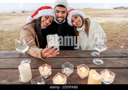 Three mixed race friends with lowered mask sitting on a wooden table outdoor in sea resort celebrating Christmas and New Year event making selfie with Stock Photo
