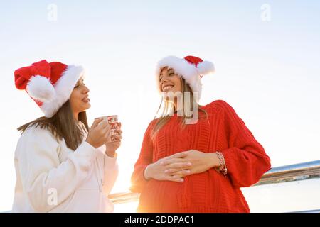 Two women in Santa Claus hat having bonding outdoor holding a Christmas tea mug - Bottom view of beautiful woman leaning to a metal railing talking to Stock Photo