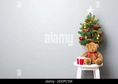 Gift boxes, teddy bear and small decorated Christmas tree on stool chair in gray wall background with copy space Stock Photo