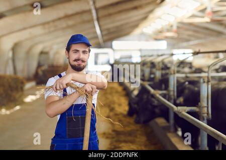 Young farm worker leaning on spade and looking at camera, standing in barn with feeding cattle Stock Photo