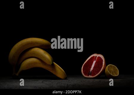 Three ripe bananas lie on top of each other on dark gray concrete, opposite lemon and grapefruit. Low key. Stock Photo