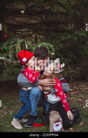 Father in red Christmas hat and two daughters in red sweaters happy near Christmas tree outdoor in the yard of the house before holidays, europe Stock Photo