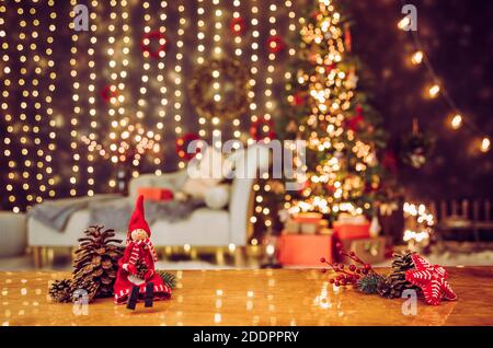 Selective focus on red elf figurine with pinecones and felt star Christmas ornament. On blurred bokeh background is decorated living room with sofa. Stock Photo