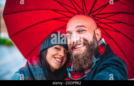 happy couple of a bearded bald man and a caucasian brunette woman taking a selfie portrait holding an umbrella during christmas celebration holidays. Stock Photo