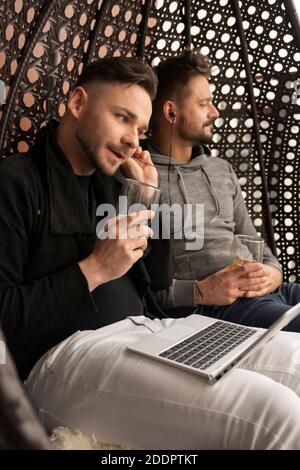 Family time on the balcony. Young handsome gay couple listening to the music on the headphones and relaxing on the swing with beer . High quality photo Stock Photo