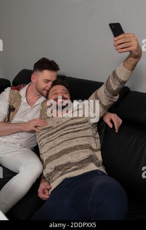 Male romantic gay couple relaxing on the couch. Boyfriend kissing his partner in the head. Taking selfie with smartphone. High quality photo Stock Photo