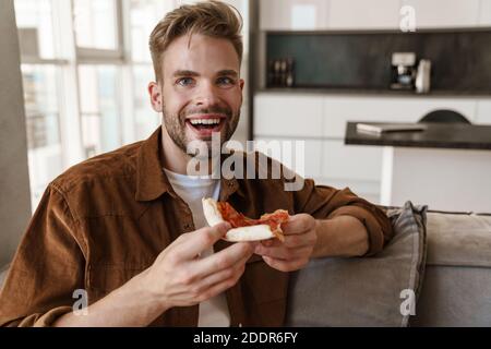 Photo of happy cheery handsome young man sitting on sofa indoors at home and eating pizza Stock Photo