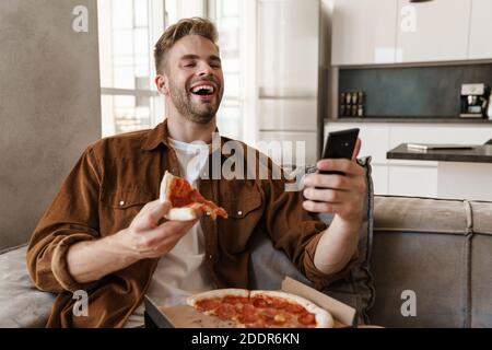 Image of laughing handsome young man sitting on sofa indoors at home and eating pizza while using mobile phone Stock Photo