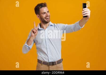 Photo of smiling man gesturing peace sign while taking selfie on cellphone isolated over yellow background Stock Photo