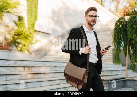 Handsome unshaven businessman in earphones using cellphone while walking down stairs on city street Stock Photo