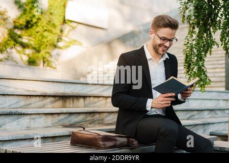 Handsome smiling businessman in earphones reading book while sitting on bench near stairs outdoors Stock Photo
