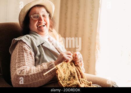 Elderly woman laughs out loud while knitting sweater for her grandchildren. Stock Photo