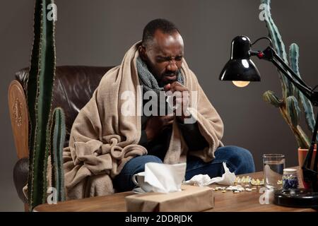 Runny nose. Young man wrapped in a plaid looks sick, ill, sneezing and coughing sitting on armchair at home. Healthcare and medicine, ill prevention, seasonal illness symptoms and self-protection. Stock Photo