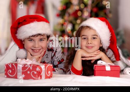 Two cute children, a girl and a boy, in santa claus hats, cuddle on white bed Stock Photo