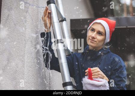 woman with santa hat standing on ladder outdoors and installing christmas lights and decors on house facade Stock Photo