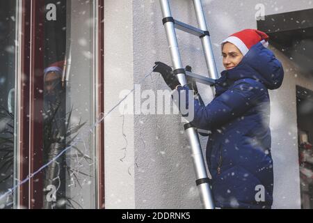 woman with santa hat standing on ladder and decorating house exterior with outdoor christmas string lights. snowing Stock Photo
