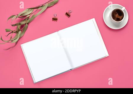 Empty open notebook, two golden binder clips, cup of coffee and green twig on pink background. Stationery, supplies for office. Close up, copy space Stock Photo