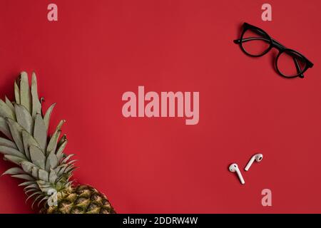 Stylish glasses with black frames, wireless earphones or earbuds and ripe pineapple against red studio background. Close up, copy space Stock Photo