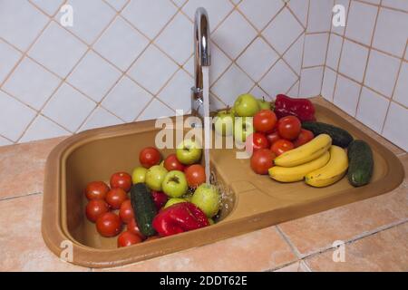 The jet of water washes fruits and vegetables in the sink of the rustic kitchen of a home with a white tile background. Stock Photo