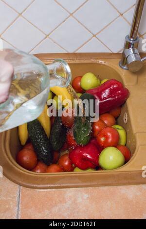 Stream of water on fruits and vegetables. Banana, cucumber, tomato, apple, and red pepper washed with water from a glass bottle in the kitchen sink. Stock Photo